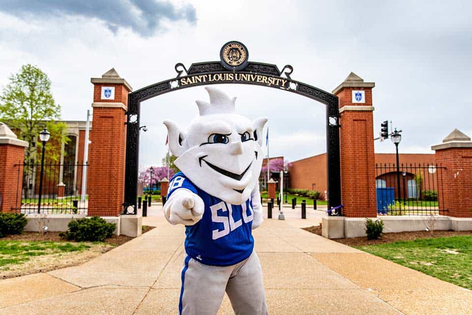 The Billiken mascot points at the camera. The Saint Louis University pedastrian gate is in the background.