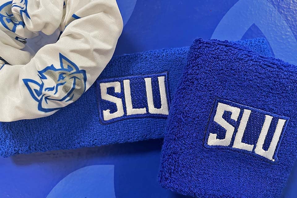 A scrunchie with small Billiken head logos printed on it sits on a table next to a head and wrist band that each say SLU.