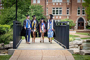 Four students wearing graducation and gowns walk across campus.