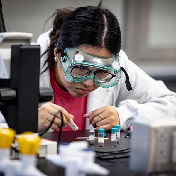 A female student wearing a white lab coat and safety glasses examines specimens in small glass tubes while working in a lab.