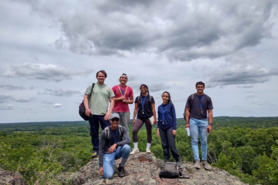 A group of six students stand and kneel at the top of a rocky bluff overlooking the Ozark forests. 