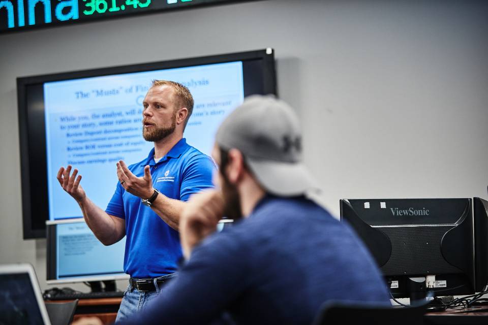 Business Professor lectures at the front of a room in front of a screen and a stock ticker.