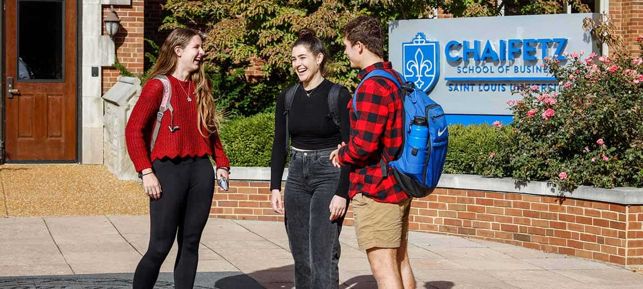 Three students chat and laugh outside Cook Hall. A large sign reading “Chaifetz School of Business” can be seen behind them.