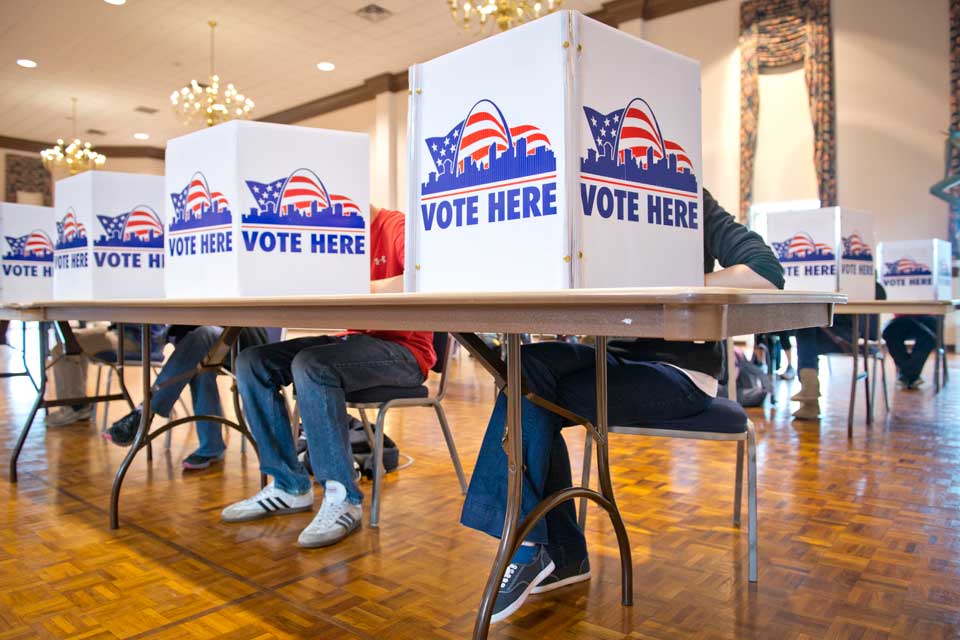 Students vote behind small barriers that read Vote Here at tables in the Saint Louis Room.