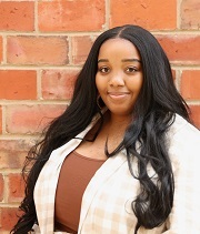 Aubra Ladd, headshot, stands in front of a brick wall and smiles. She is wearing a tan and white plaid shirt over a brown top.