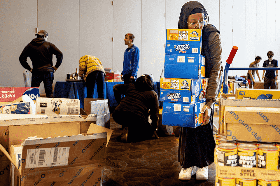 A student carries several large boxes of Frosted Flakes cereal while others work around her.