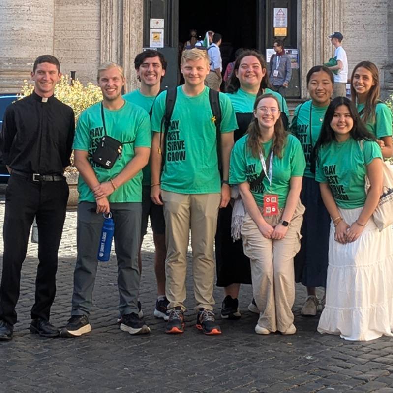 A group of students stands on a street in a rome with a priest. The students wear shirts that say pray, rest, serve, grow.