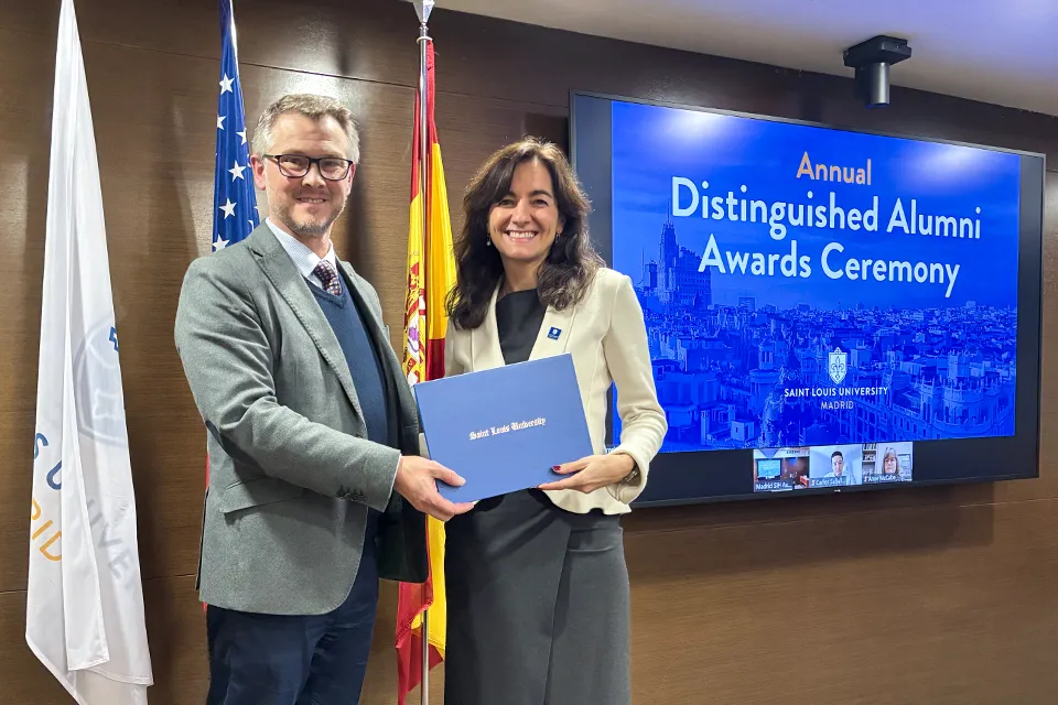 A man hands a certificate to a woman on stage, with some flags and a large screen behind them that reads Annual Distinguished Alumni Awards Ceremony