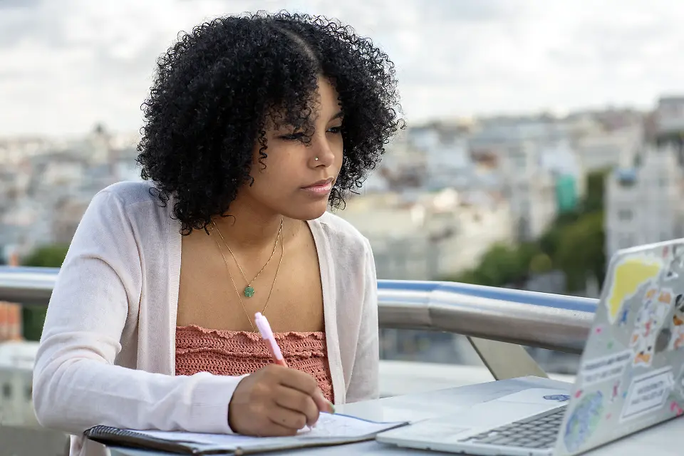 Student taking notes on a notepad and looking at a laptop while sitting at a table on an outdoor balcony.