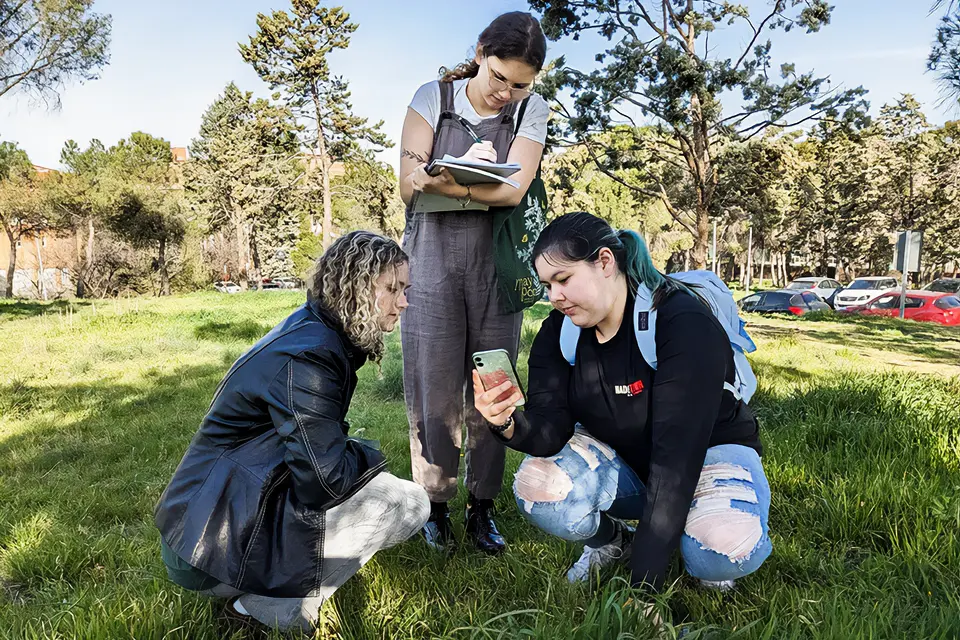 Two students kneel on the ground while a third stands and records information in a notebook.