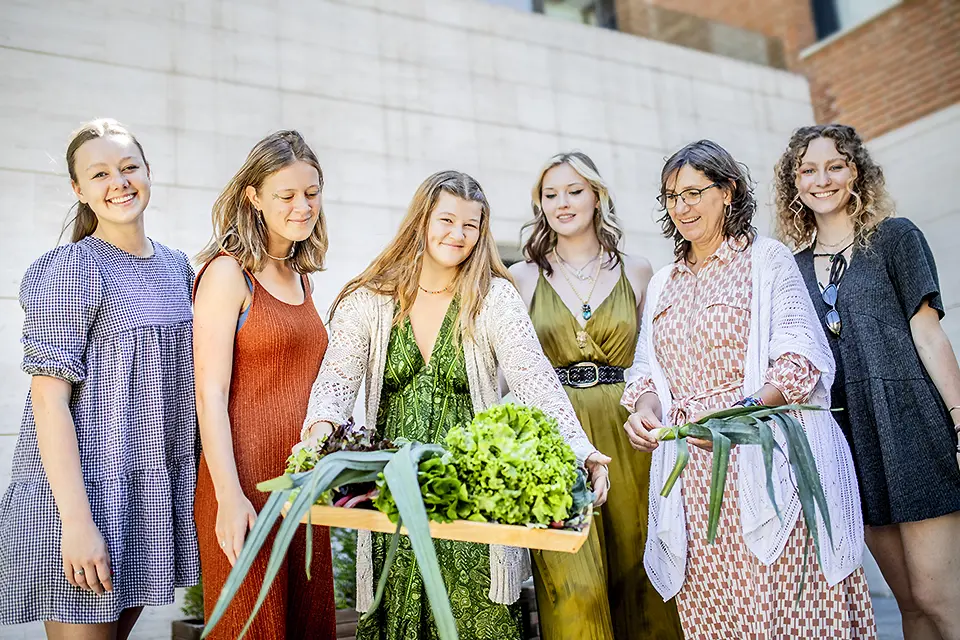 A student holds a large wooden tray covered in lettuce and other vegetables while several others look on.