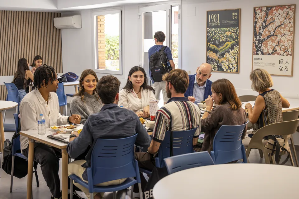 President Feser Visits SLU-Madrid in His First 100 Days Eight people sit at a table in a cafeteria space where there are decorative posters on the wall and behind them there is a door that leads to a patio.