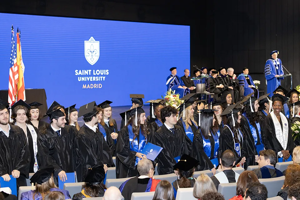 Students in graduation attire face audience with stage in background.