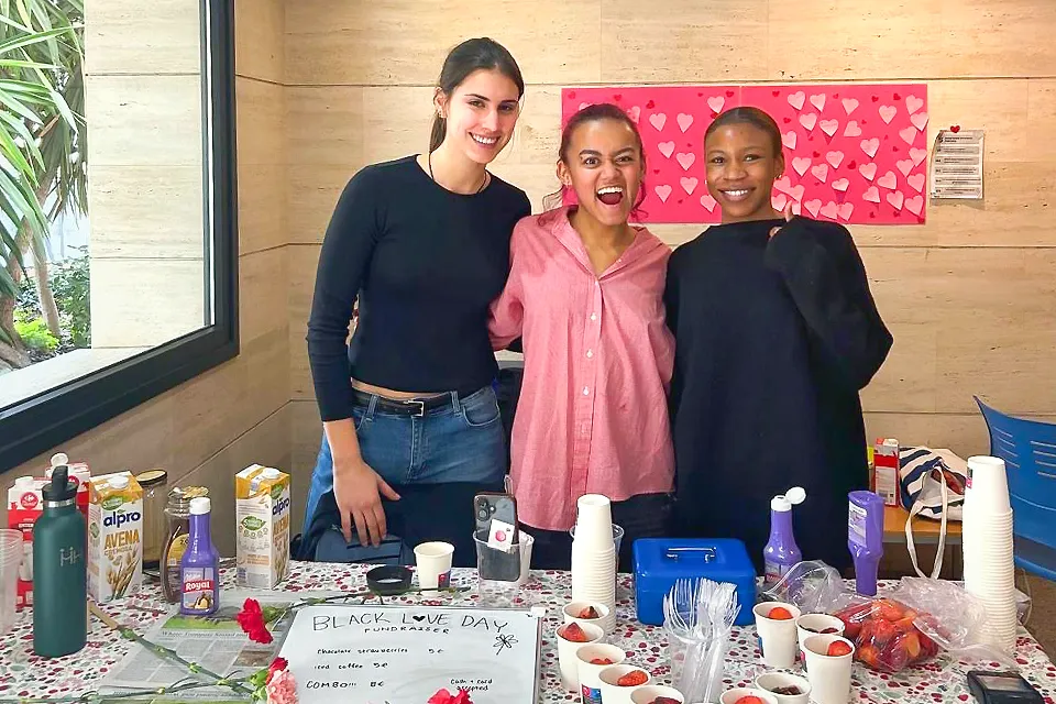 Three students stand smiling behind a table at a "Black Love Day" fundraiser event. The table is decorated with a heart-themed backdrop and holds cups of strawberries, chocolate syrup, milk cartons, flowers, and a handwritten sign reading "Black Love Day Fundraiser."