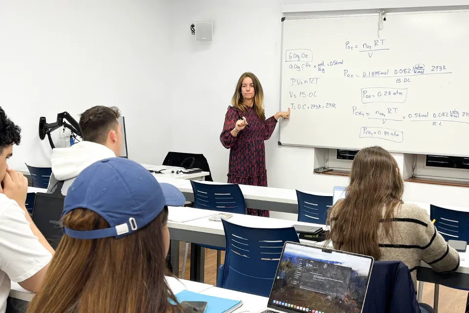 Tania de la Fuente, Ph.D., teaching a chemistry class in a Padre Rubio Hall classroom. Woman in front of class while pointing to an equation on the whiteboard behind her with a computer monitor next to her while students look toward the front of the room.