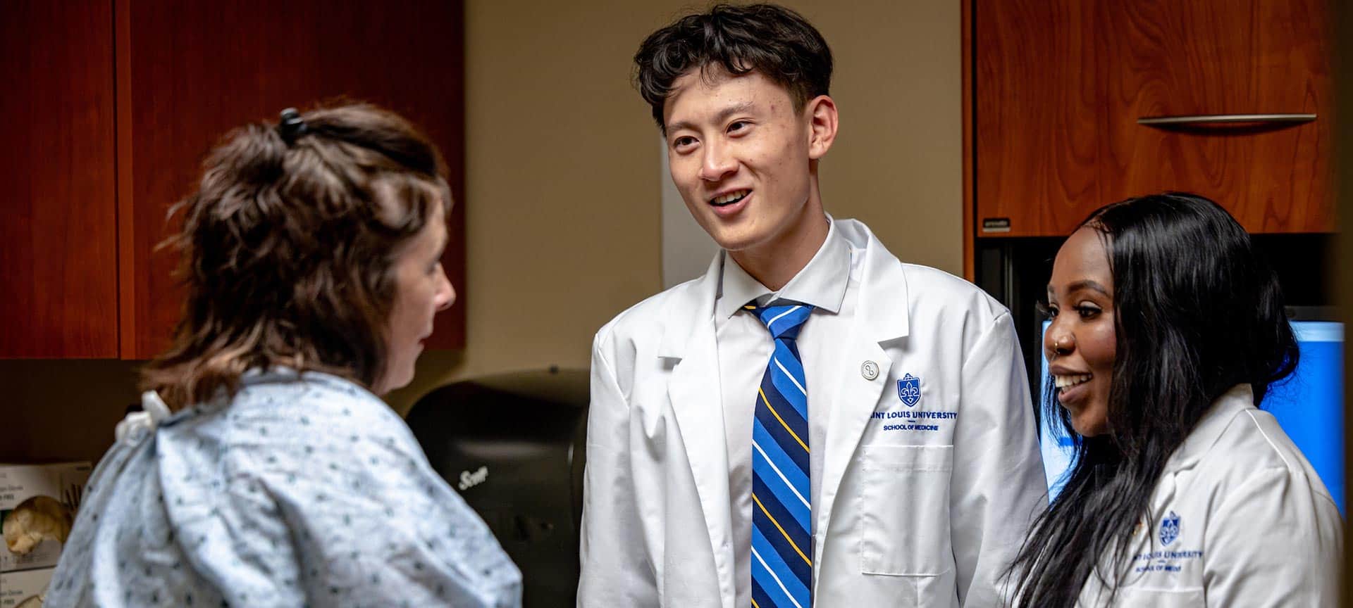 Two medical students in white coats visiting with a patient in an exam room