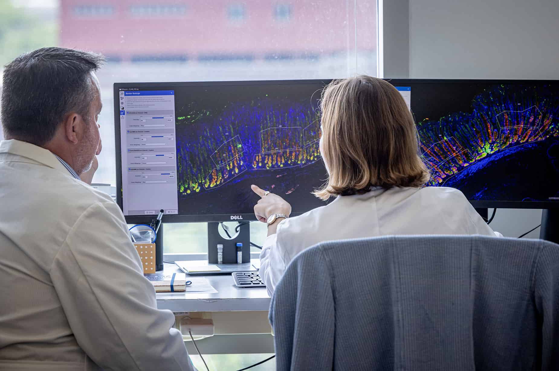 A student sits at a desk with a large computer monitor displaying research information. A mentor sits to the side watching her work.