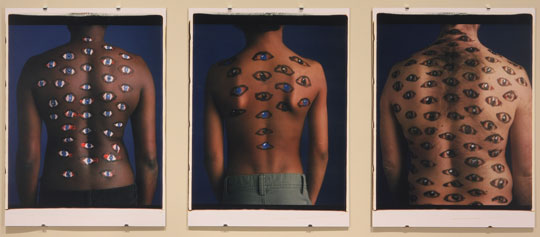 An artwork by Maria Magdalena Campos-Pons titled, Sagrada Familia (Holy Family). Three photographs depict, from left to right, the back of a woman, of a young boy, and an adult man. They have varying skin tones and a variety of stylized eyes painted on their backs.