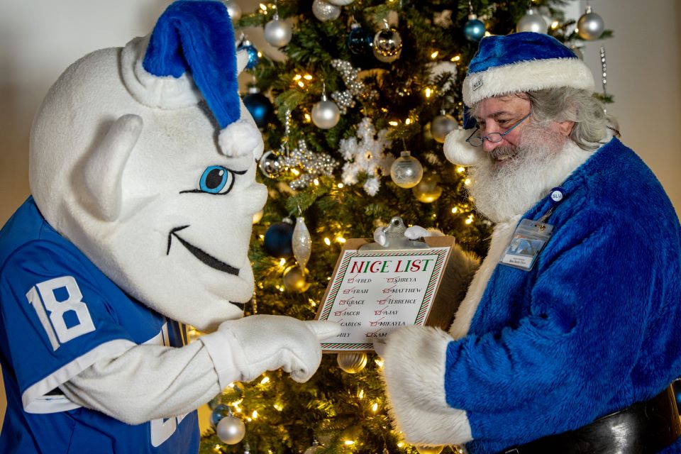 Billiken points to a nice list on a clipboard held by Santa Clause in a blue suit in front of a Christmas tree. 
