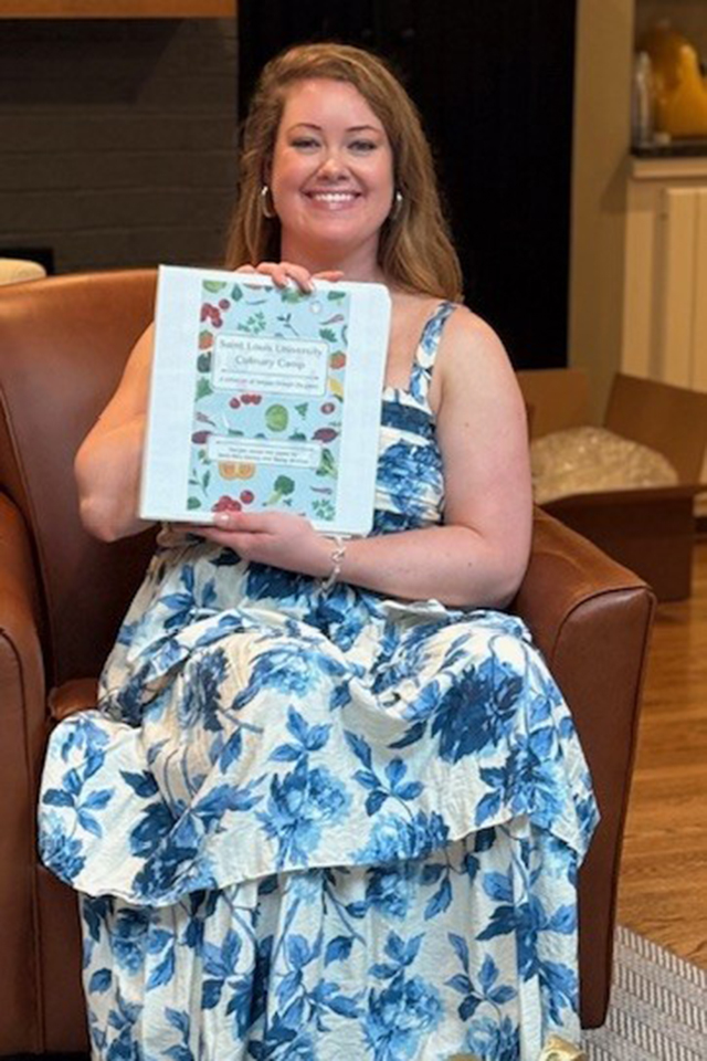 A woman in a blue-patterned dress sits in a chair. She holds a recipe book and smiles for a photo indoors.