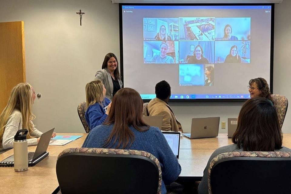 Interior of a classroom. An instructor stands at the front while students sit at desks. A screen with videoconference participants is in the background.