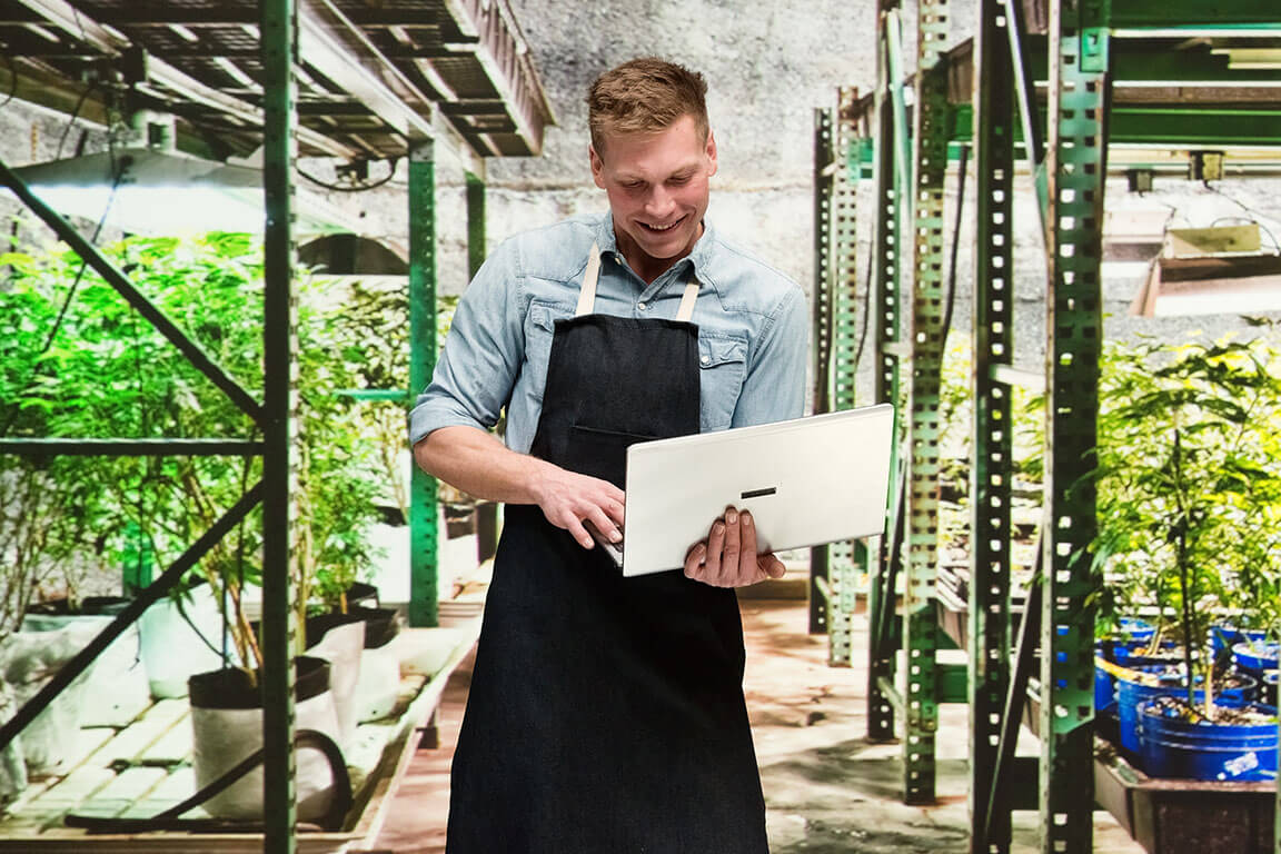 A man in an apron standing in a greenhouse holding a laptop