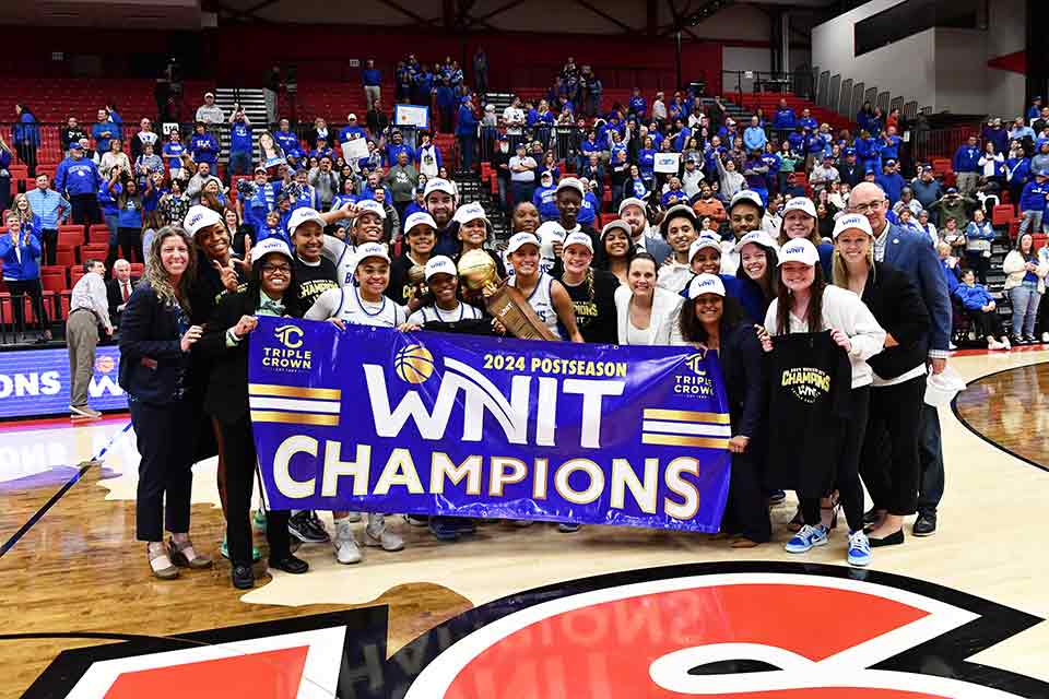 SLU's women's basketball team stand in the middle of a basketball court holding a banner that reads "WNIT Champions."