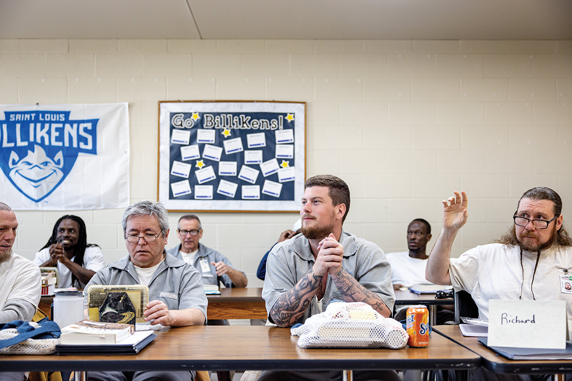 Several incarcerated men of various ages and races sit at long tables in a prison classroom. One types on a tablet. Another raises his hand. 